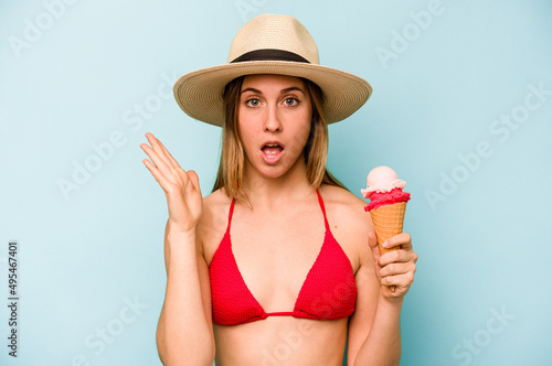Young caucasian woman wearing a bikini and holding an ice cream isolated on blue background surprised and shocked.