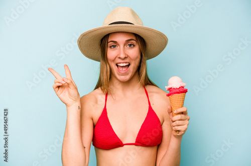 Young caucasian woman wearing a bikini and holding an ice cream isolated on blue background joyful and carefree showing a peace symbol with fingers.