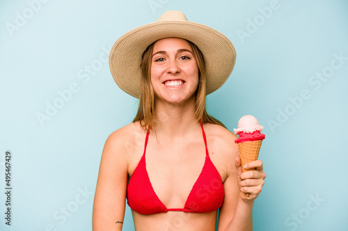 Young caucasian woman wearing a bikini and holding an ice cream isolated on blue background laughing and having fun.