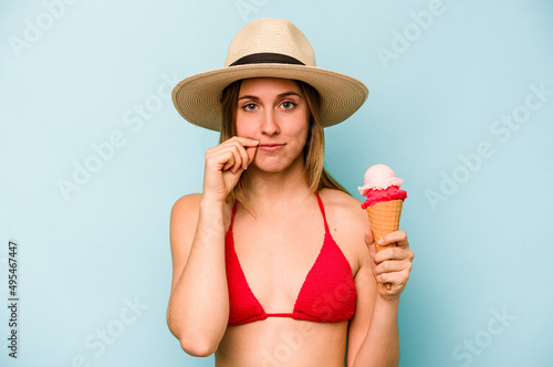 Young caucasian woman wearing a bikini and holding an ice cream isolated on blue background with fingers on lips keeping a secret.