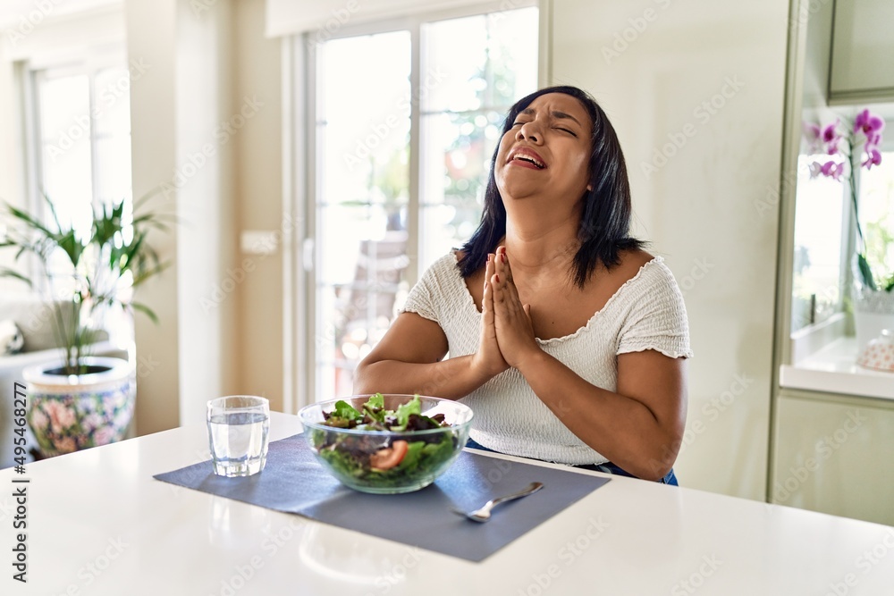 Young hispanic woman eating healthy salad at home begging and praying with hands together with hope expression on face very emotional and worried. begging. Young hispanic woman eating healthy salad at home begging and praying with hands together with hope expression on face very emotional and worried. begging.