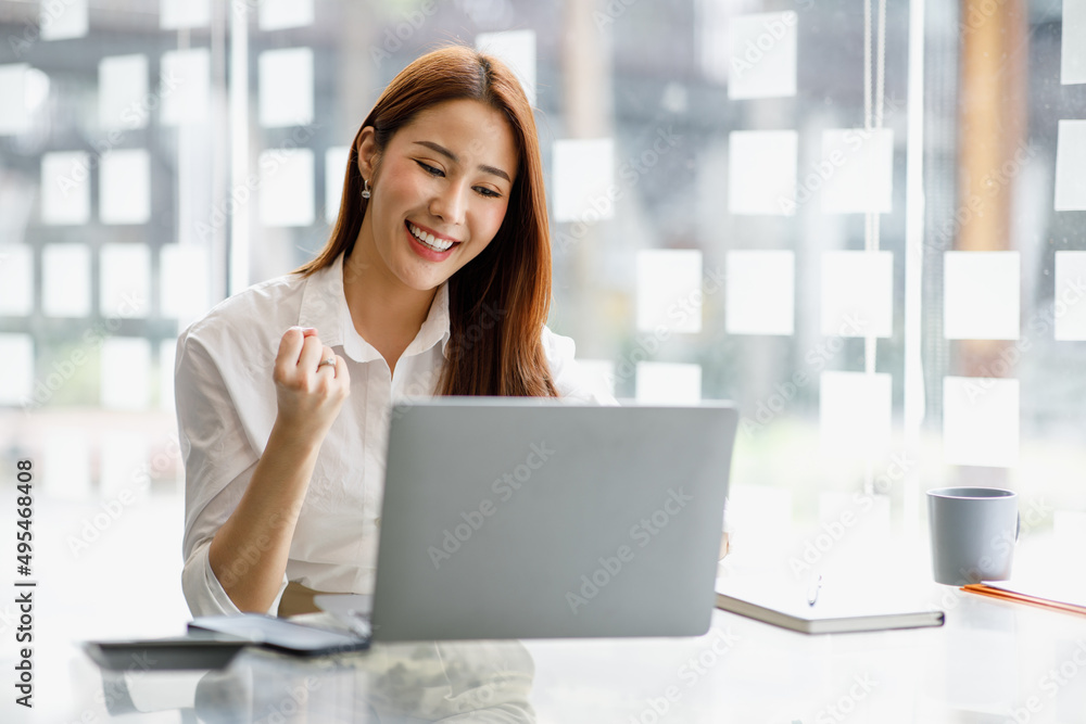 Yes! Happy excited asian woman at workstation triumphing with laptop computer raised hands Yes! Happy excited asian woman at workstation triumphing with laptop computer raised hands