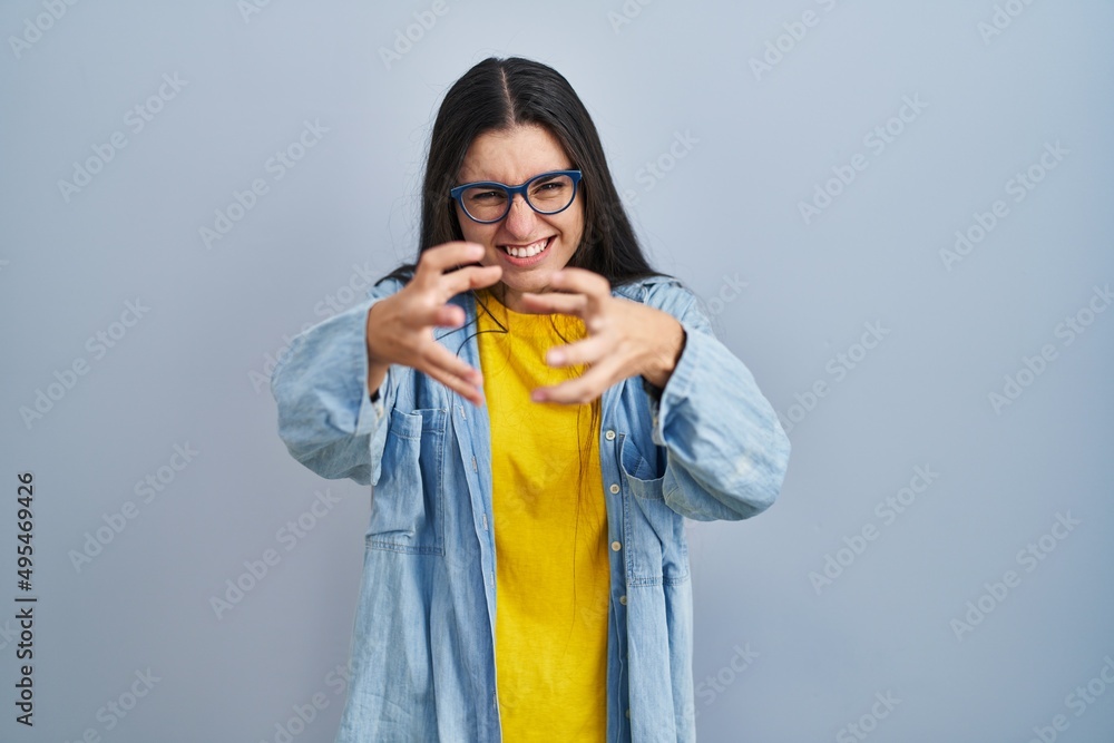 Young hispanic woman standing over blue background shouting frustrated with rage, hands trying to strangle, yelling mad Young hispanic woman standing over blue background shouting frustrated with rage, hands trying to strangle, yelling mad