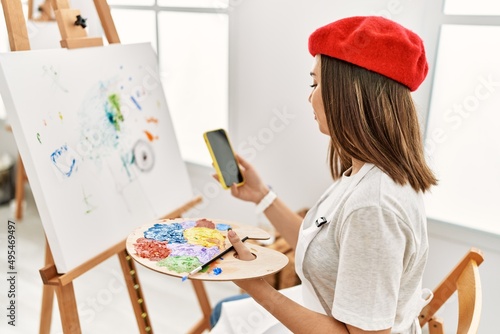 Young hispanic artist woman wearing french beret drawing using smartphone at art studio.
