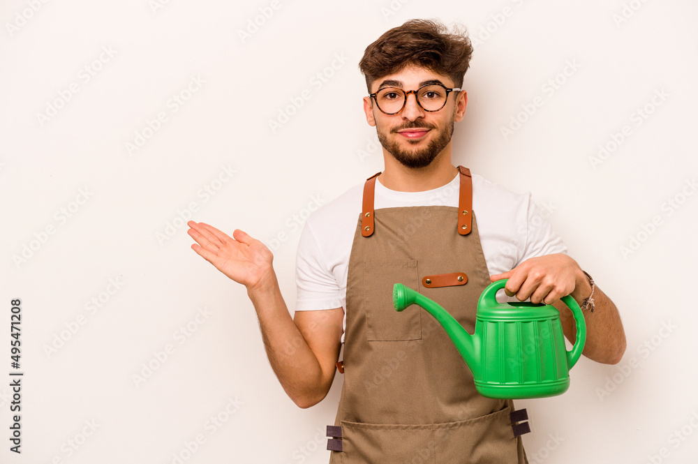 Young gardener hispanic man holding a watering can isolated on white background showing a copy space on a palm and holding another hand on waist. Young gardener hispanic man holding a watering can isolated on white background showing a copy space on a palm and holding another hand on waist.