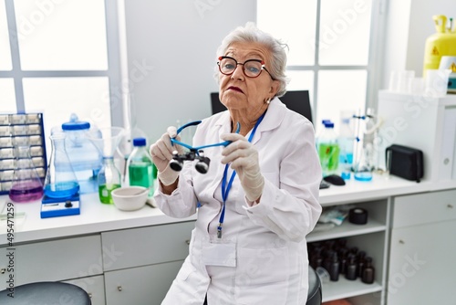 Senior woman with grey hair working at scientist laboratory using magnifying glasses clueless and confused expression. doubt concept.