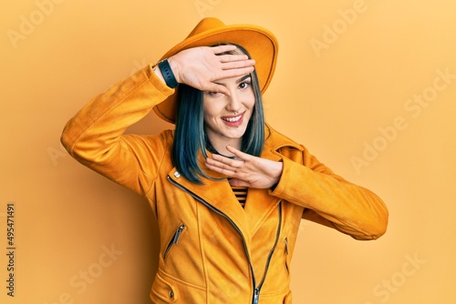 Young modern girl wearing yellow hat and leather jacket smiling cheerful playing peek a boo with hands showing face. surprised and exited