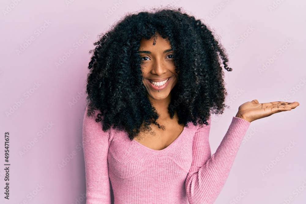 African american woman with afro hair wearing casual pink shirt smiling cheerful presenting and pointing with palm of hand looking at the camera. African american woman with afro hair wearing casual pink shirt smiling cheerful presenting and pointing with palm of hand looking at the camera.