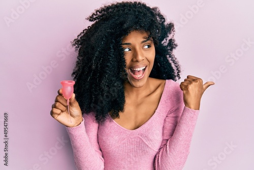 African american woman with afro hair holding menstrual cup pointing thumb up to the side smiling happy with open mouth