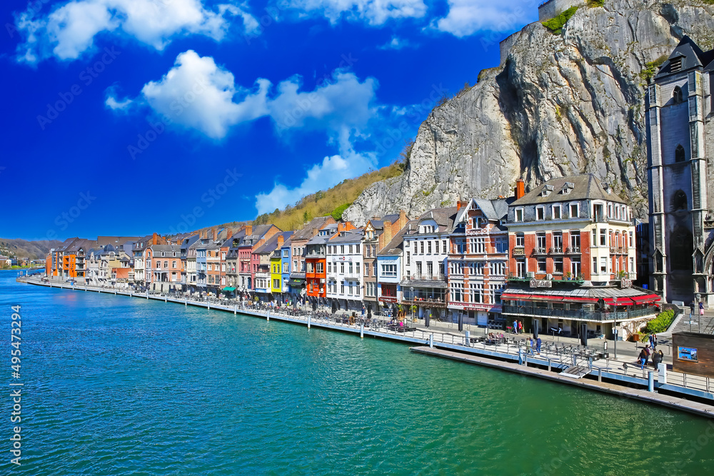 Dinant, Belgium - March 9. 2022: Idyllic colorful belgian town, gothic church, steep rock face wall, waterfront promenade, river meuse with cafes and restaurants