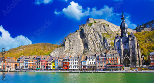 Dinant, Belgium - March 9. 2022: View over river meuse on picturesque series of old colorful houses, rock wall with citadel, gothic church against clear blue winter sky, fluffy clouds