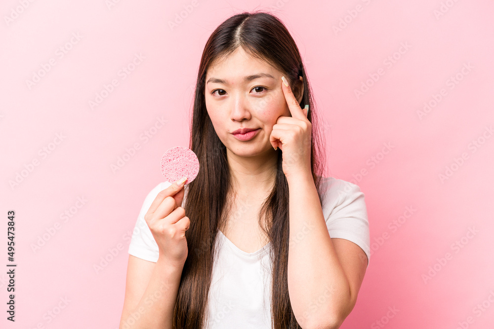 Young asian woman holding facial sponge isolated on pink background pointing temple with finger, thinking, focused on a task. Young asian woman holding facial sponge isolated on pink background pointing temple with finger, thinking, focused on a task.