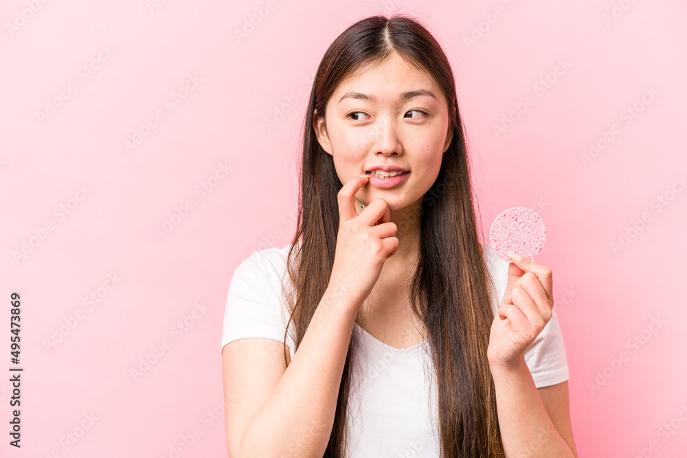 Young asian woman holding facial sponge isolated on pink background relaxed thinking about something looking at a copy space. Young asian woman holding facial sponge isolated on pink background relaxed thinking about something looking at a copy space.