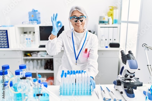 Middle age grey-haired woman wearing scientist uniform doing ok sign with fingers, smiling friendly gesturing excellent symbol