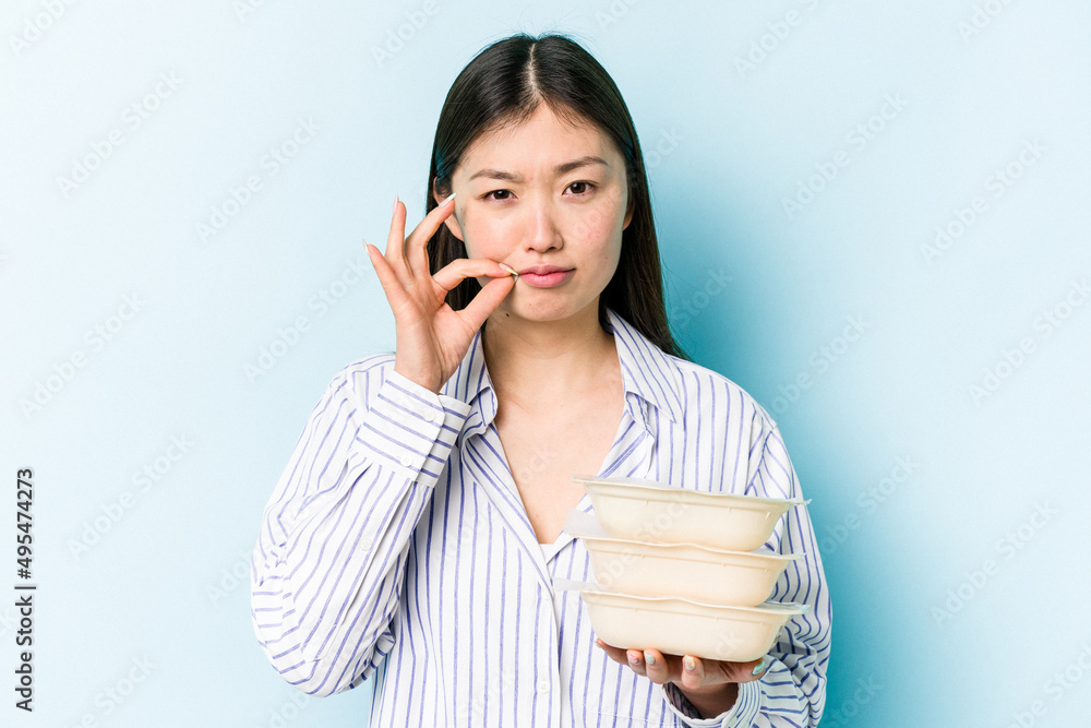 Young asian woman holding Tupperware isolated on blue background with fingers on lips keeping a secret. Young asian woman holding Tupperware isolated on blue background with fingers on lips keeping a secret.