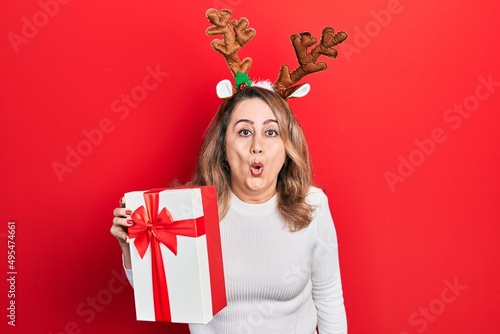 Middle age caucasian woman wearing cute christmas reindeer horns holding gift scared and amazed with open mouth for surprise, disbelief face