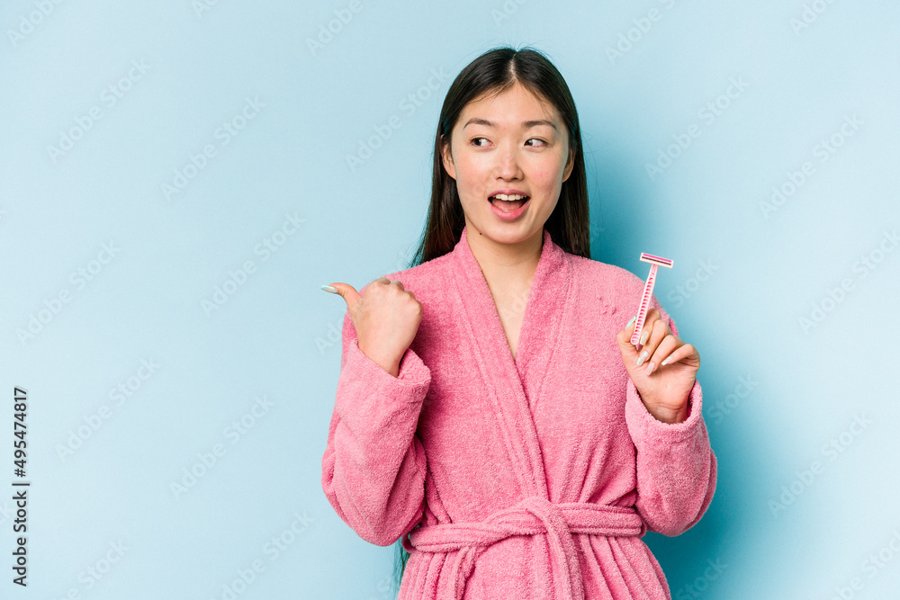Young asian woman holding razor blade isolated on blue background points with thumb finger away, laughing and carefree. Young asian woman holding razor blade isolated on blue background points with thumb finger away, laughing and carefree.