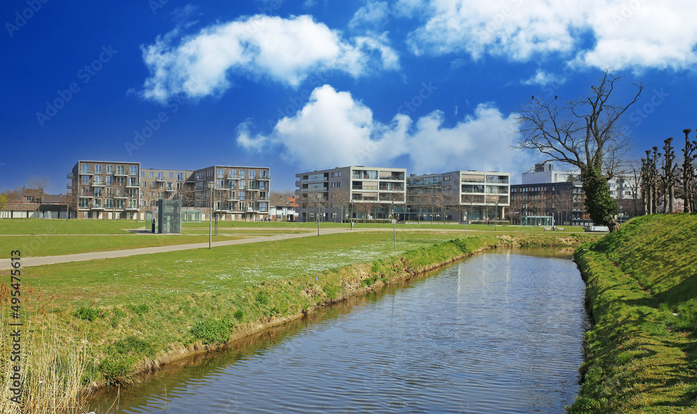 Sittard, Netherlands - March 25. 2022: View beyond water canal over green grass lawn on series of modern residential homes, blue sky with fluffy clouds