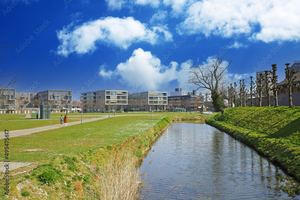 Sittard, Netherlands - March 25. 2022: View beyond water canal over green grass lawn on series of modern residential homes, blue sky with fluffy clouds