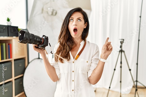 Beautiful caucasian woman working as photographer at photography studio amazed and surprised looking up and pointing with fingers and raised arms.