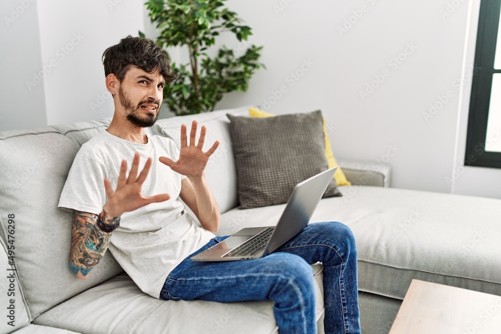 Hispanic man with beard sitting on the sofa afraid and terrified with fear expression stop gesture with hands, shouting in shock. panic concept.