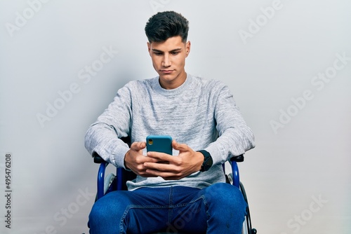 Young hispanic man sitting on wheelchair using smartphone relaxed with serious expression on face. simple and natural looking at the camera. photo