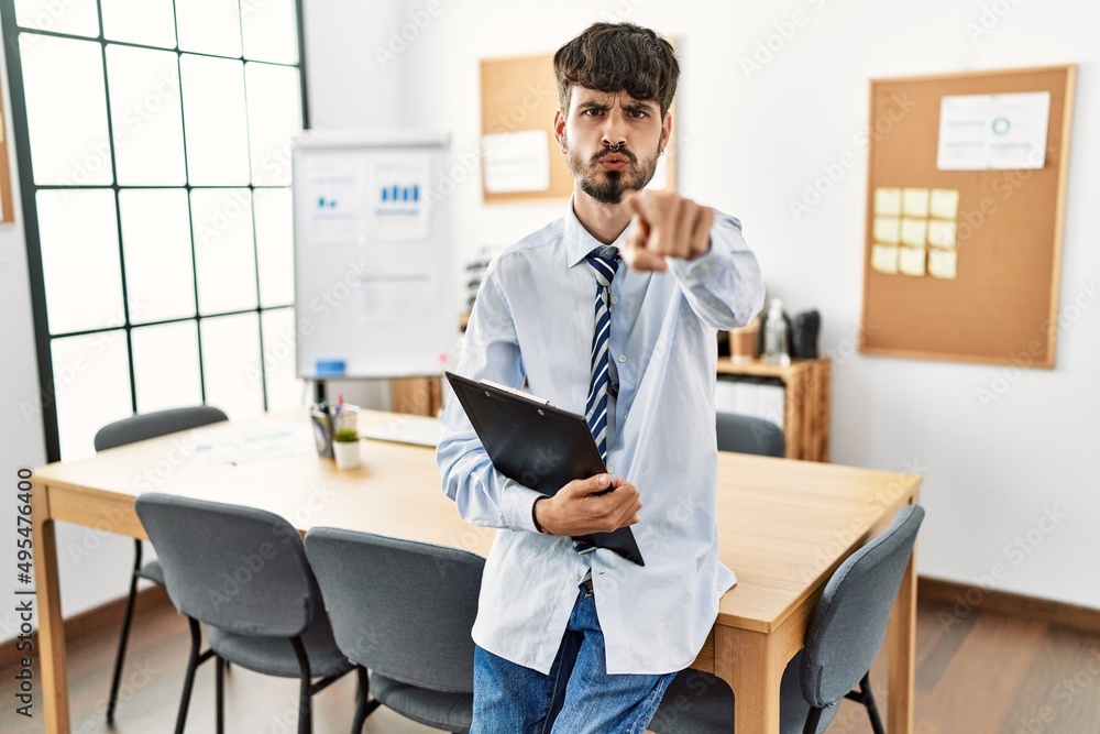 Hispanic man with beard wearing business style sitting on desk at office pointing with finger to the camera and to you, confident gesture looking serious Hispanic man with beard wearing business style sitting on desk at office pointing with finger to the camera and to you, confident gesture looking serious