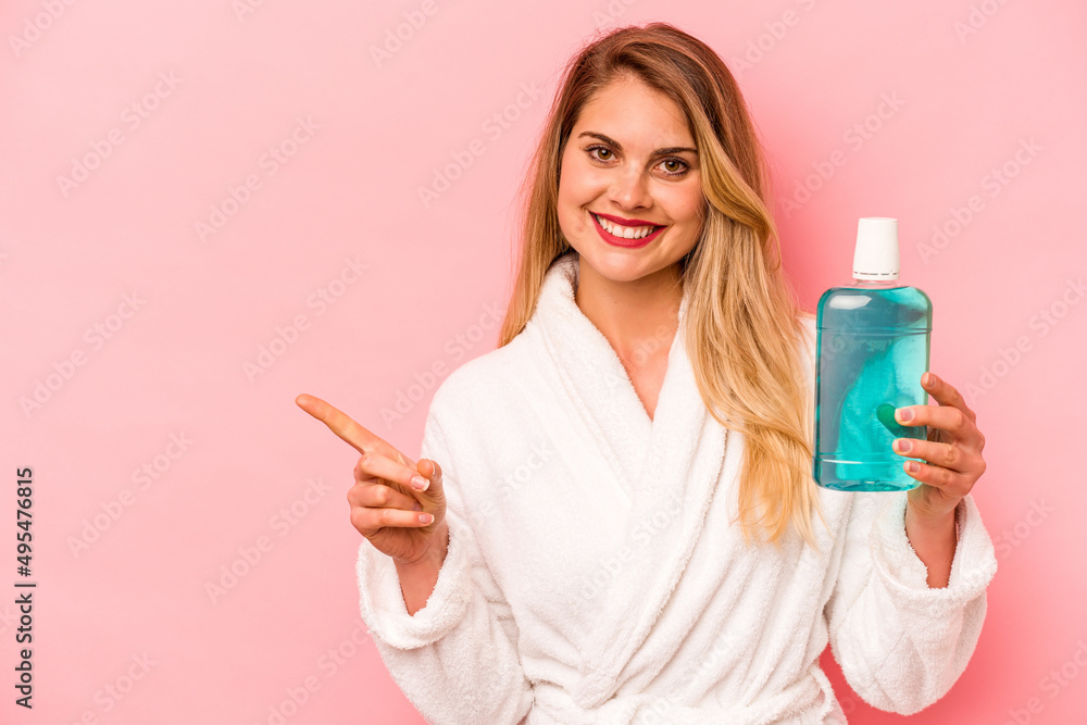 Young caucasian woman holding mouthwash wearing bathrobe isolated on pink background smiling and pointing aside, showing something at blank space. Young caucasian woman holding mouthwash wearing bathrobe isolated on pink background smiling and pointing aside, showing something at blank space.