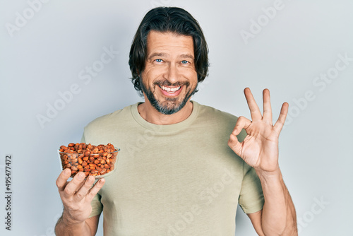 Canvas Print Middle age caucasian man holding peanuts doing ok sign with fingers, smiling fri