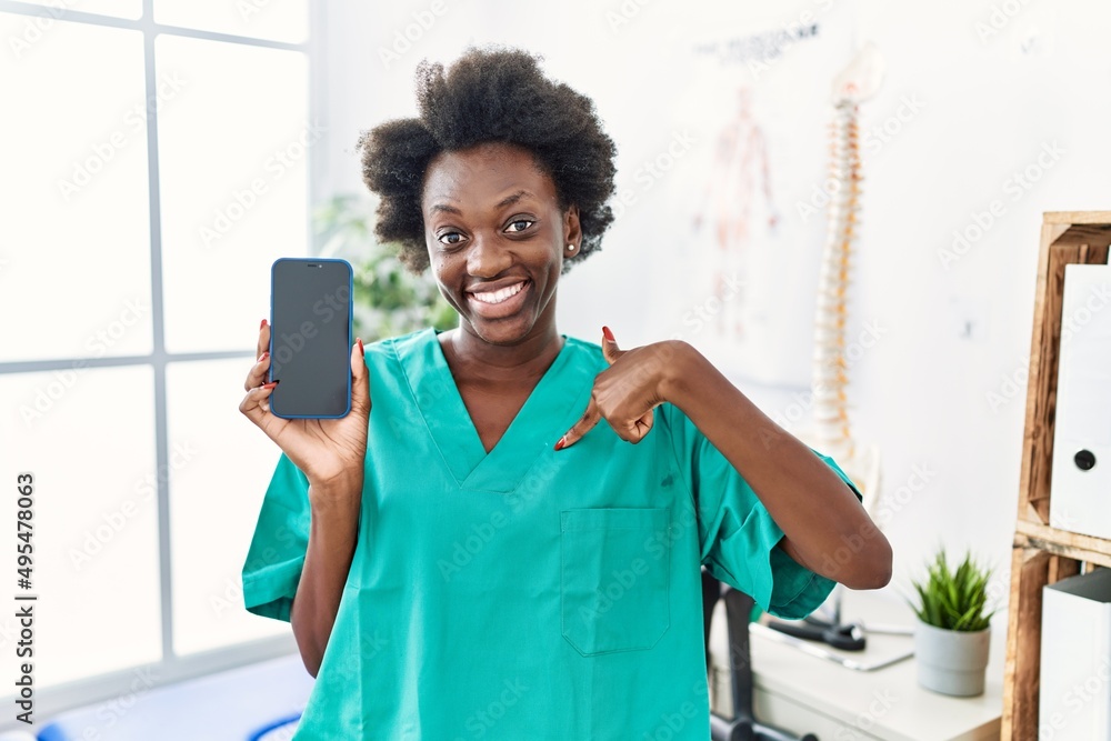 African young physiotherapist woman working at pain recovery clinic showing smartphone screen pointing finger to one self smiling happy and proud African young physiotherapist woman working at pain recovery clinic showing smartphone screen pointing finger to one self smiling happy and proud