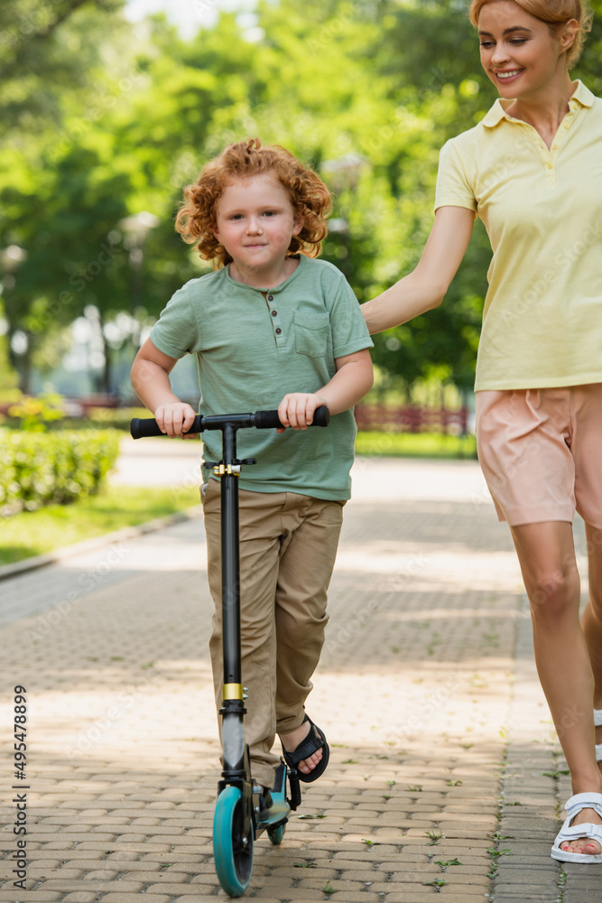 redhead boy looking at camera while riding kick scooter near smiling mom. redhead boy looking at camera while riding kick scooter near smiling mom.