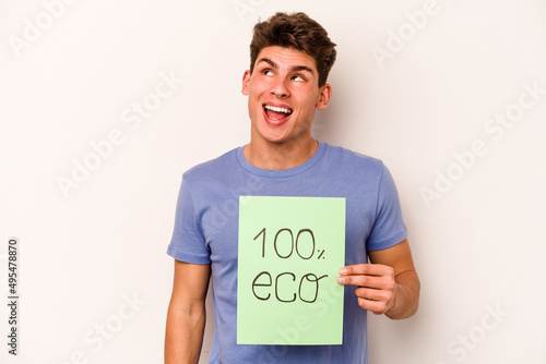 Young caucasian man holding 100% eco placard isolated on white background dreaming of achieving goals and purposes