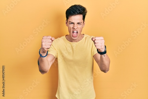 Young hispanic man wearing casual yellow t shirt angry and mad raising fists frustrated and furious while shouting with anger. rage and aggressive concept.