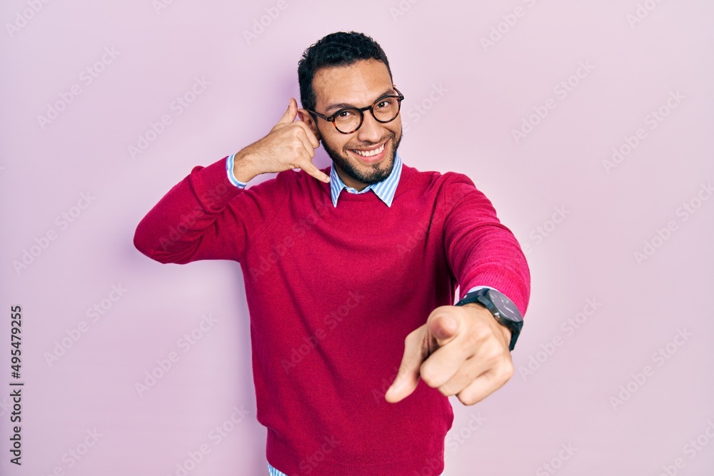 Hispanic man with beard wearing business shirt and glasses smiling doing talking on the telephone gesture and pointing to you. call me. Hispanic man with beard wearing business shirt and glasses smiling doing talking on the telephone gesture and pointing to you. call me.