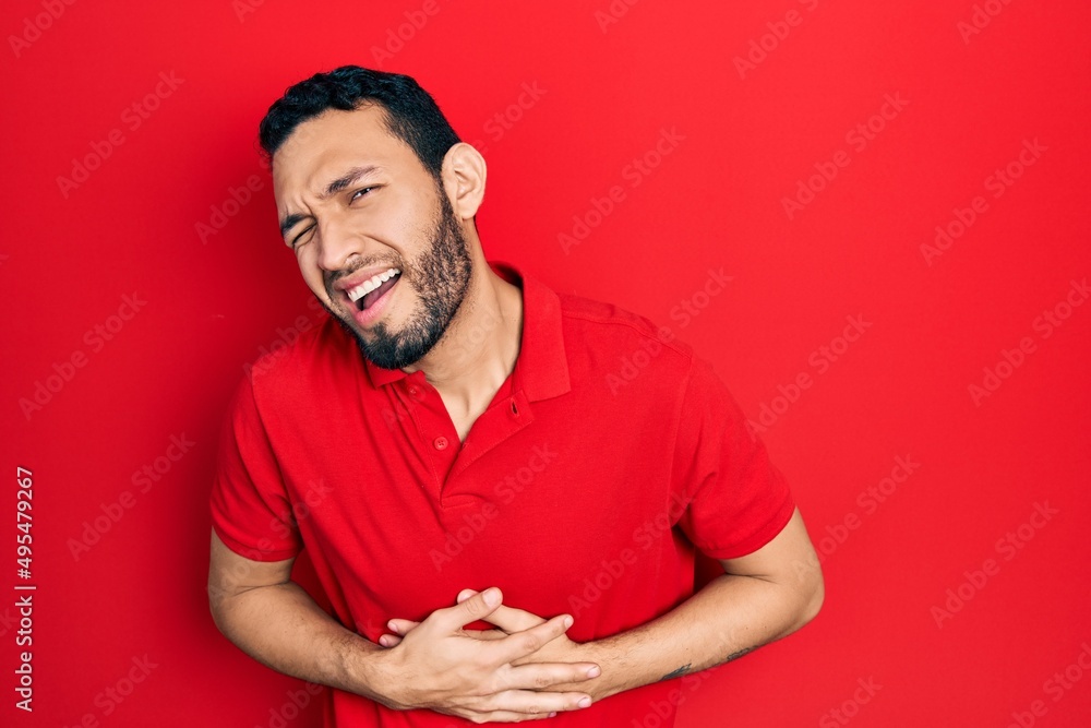 Hispanic man with beard wearing casual red t shirt with hand on stomach because nausea, painful disease feeling unwell. ache concept. Hispanic man with beard wearing casual red t shirt with hand on stomach because nausea, painful disease feeling unwell. ache concept.