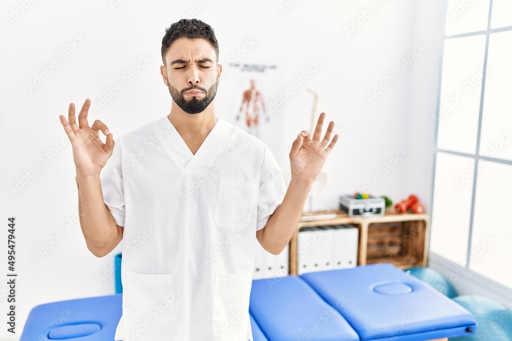 Young handsome man with beard working at pain recovery clinic relax and smiling with eyes closed doing meditation gesture with fingers. yoga concept. Young handsome man with beard working at pain recovery clinic relax and smiling with eyes closed doing meditation gesture with fingers. yoga concept.