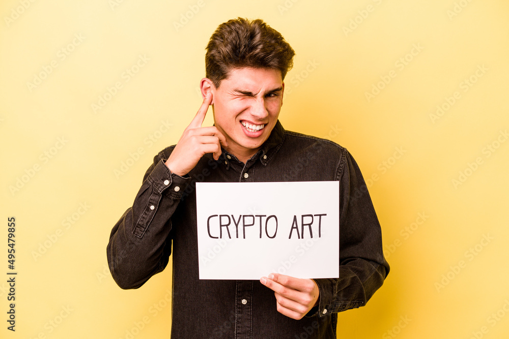 Young caucasian man holding crypto art placard isolated on yellow background covering ears with hands. Young caucasian man holding crypto art placard isolated on yellow background covering ears with hands.