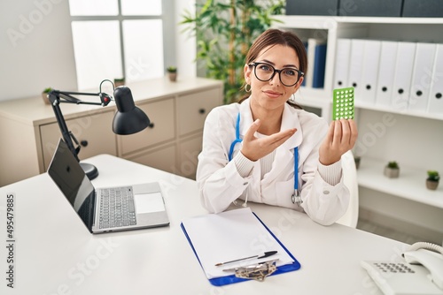 Young hispanic woman wearing doctor uniform holding birth control pills at clinic