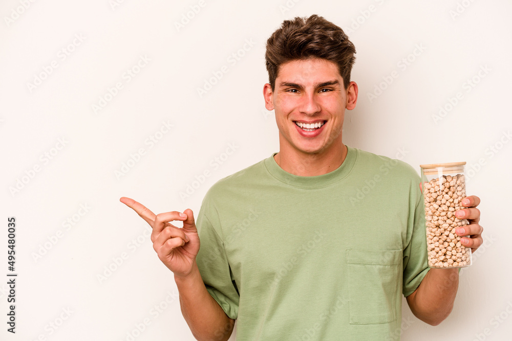 Young caucasian man holding chickpeas isolated on white background smiling and pointing aside, showing something at blank space.