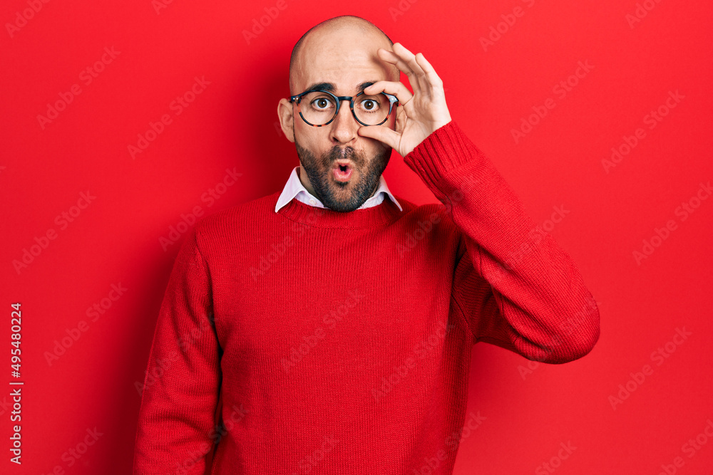 Young bald man wearing casual clothes and glasses doing ok gesture shocked with surprised face, eye looking through fingers. unbelieving expression.