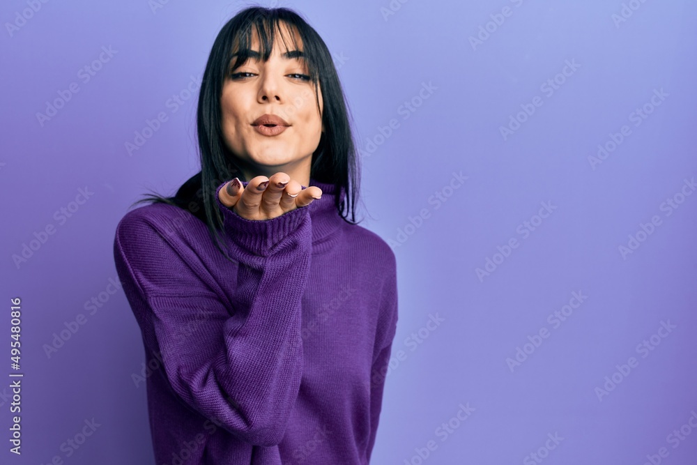 Young brunette woman with bangs wearing turtleneck sweater looking at the camera blowing a kiss with hand on air being lovely and sexy. love expression. Young brunette woman with bangs wearing turtleneck sweater looking at the camera blowing a kiss with hand on air being lovely and sexy. love expression.