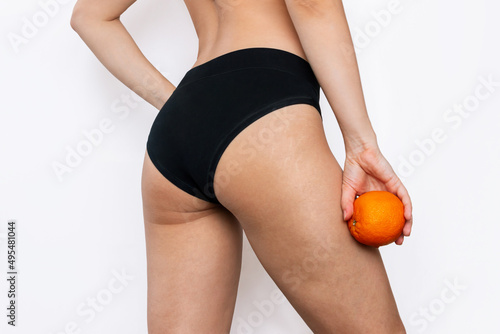 Cropped shot of young woman with cellulite and white stretch marks from a weight loss or weight gain on the thighs holding an orange in her hand isolated on a white background. Changes of the body