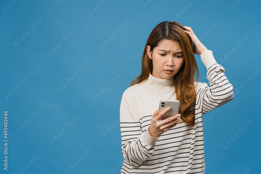 Fototapeta premium Thinking dreaming young Asia lady using phone with positive expression, dressed in casual clothing feeling happiness and stand isolated on blue background. Happy adorable glad woman rejoices success.