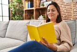Middle age hispanic woman reading book sitting on sofa at home