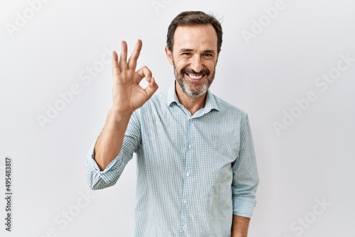 Middle age hispanic man with beard standing over isolated background smiling positive doing ok sign with hand and fingers. successful expression.