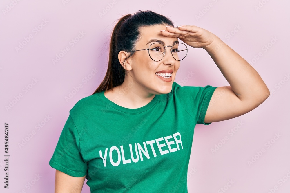 Young hispanic woman wearing volunteer t shirt very happy and smiling looking far away with hand over head. searching concept. Young hispanic woman wearing volunteer t shirt very happy and smiling looking far away with hand over head. searching concept.
