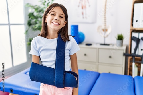 Young hispanic girl wearing arm on sling at rehabilitation clinic looking positive and happy standing and smiling with a confident smile showing teeth photo