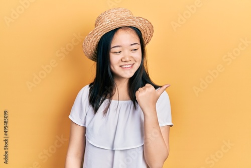 Young chinese girl wearing summer hat smiling with happy face looking and pointing to the side with thumb up.