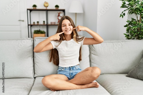Young brunette teenager sitting on the sofa at home smiling cheerful showing and pointing with fingers teeth and mouth. dental health concept.