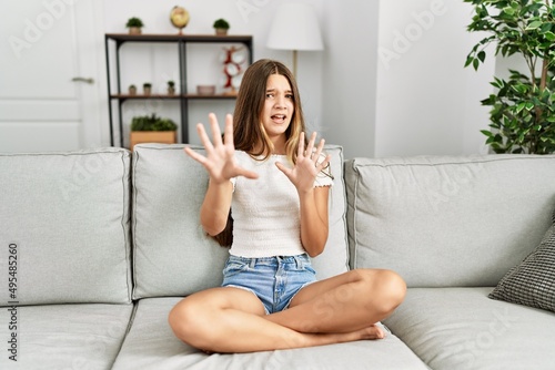 Young brunette teenager sitting on the sofa at home afraid and terrified with fear expression stop gesture with hands, shouting in shock. panic concept.